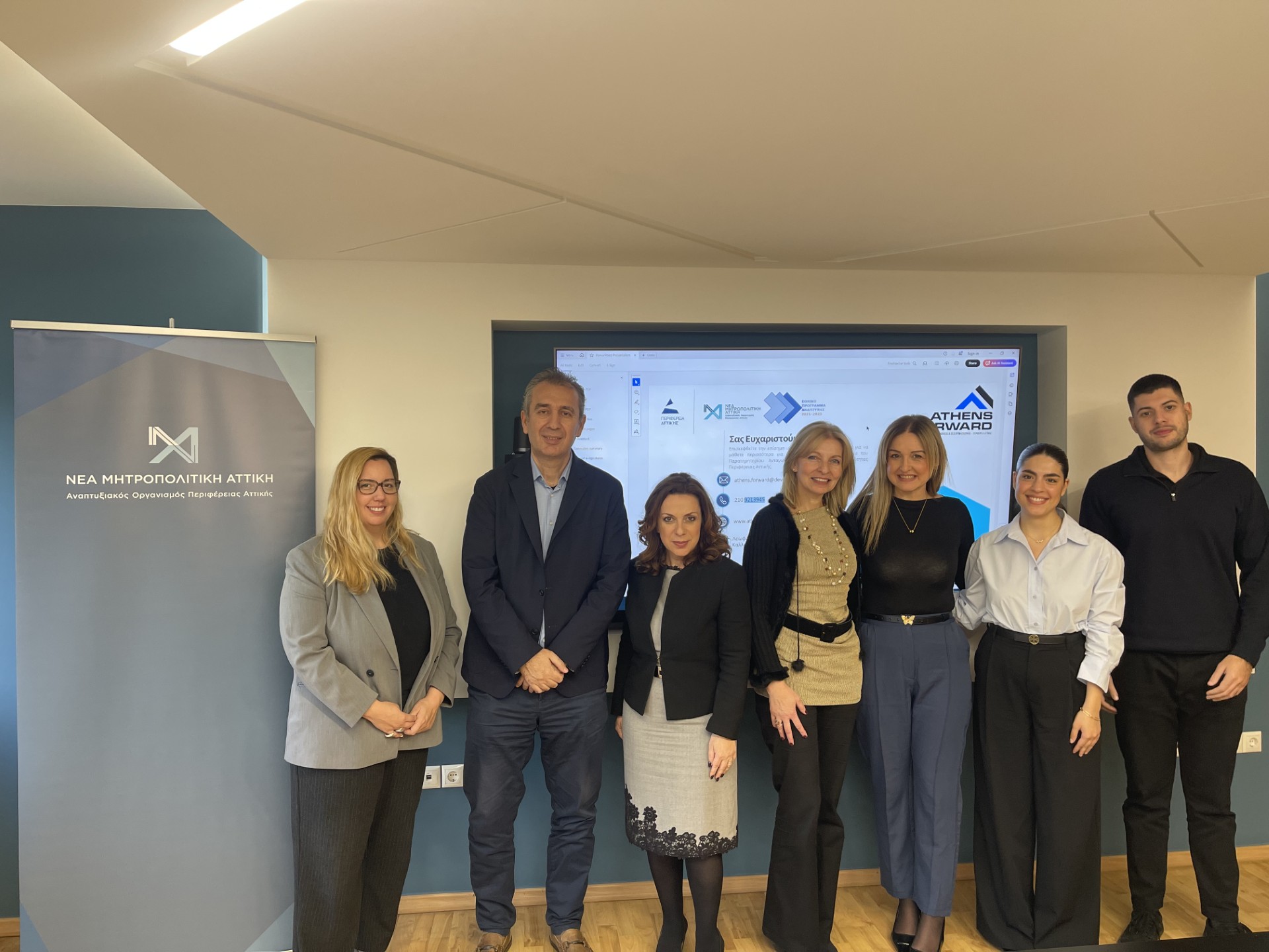 A group of six professionals poses together in an office setting, with a presentation screen and a banner in the background.